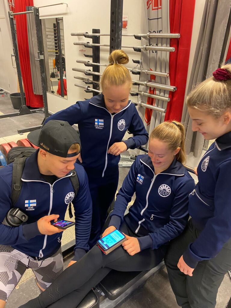 Neljä nuorta painonnostajaa käyttämässä Suomisportin mobiilisovellusta punttisalilla.