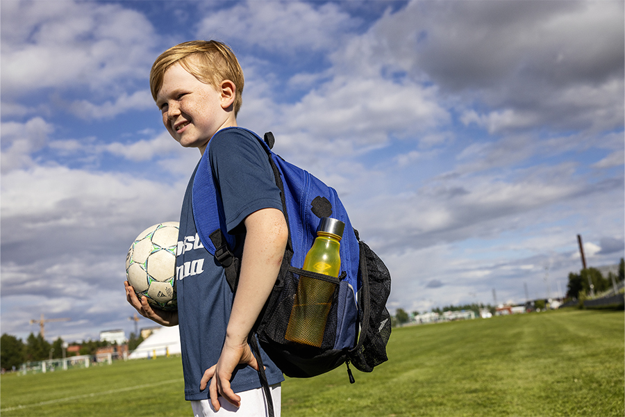 Kuvassa seisoo jalkapalloilijapoika jalkapallo sylissä ja selässään reppu, joka on paikattu mustalla vaatelaastarilla