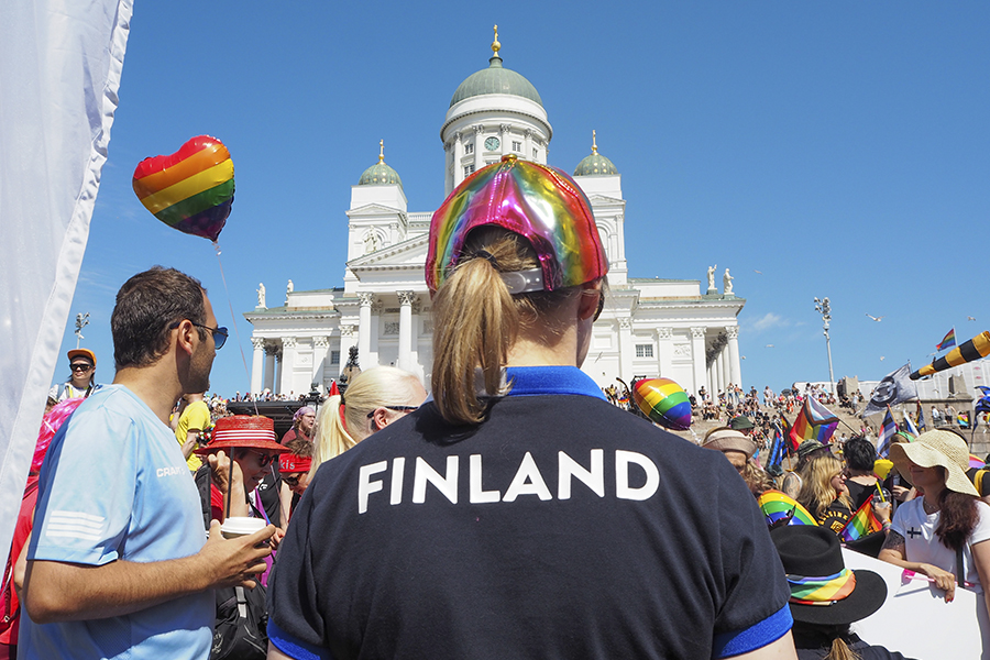 Kuvassa taustalla Senaatintorin kirkko ja etualalla seisoo nainen selin kameraan. Naisen paidan selkämyksessä lukee FINLAND ja hänellä on sateenkaaren värinen lippis päässä.