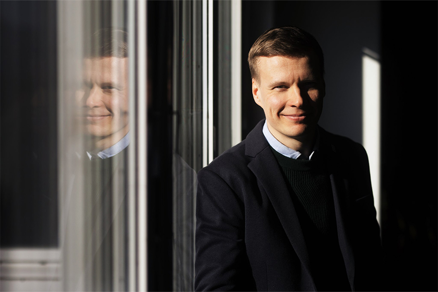 Kuvassa Matti Heikkinen nojaa ikkunaan, josta tulee kuvaan peilikuvaheijastus. Heikkinen hymyilee kuvassa.