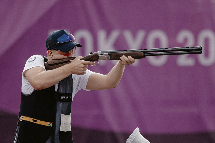 Eetu Kallioinen tähtäämässä aseellaan Tokion olympialaisissa. Kuva on otettu sivusuunnasta. Takana olevassa mainoslakanassa lukee "Tokyo 2020".