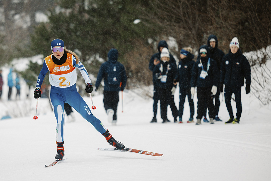 Nelli-Lotta Karppelin hiihtää vapaata kameraa kohti. Taustalla sumeana Suomen joukkueen urheilijoita kannustamassa.