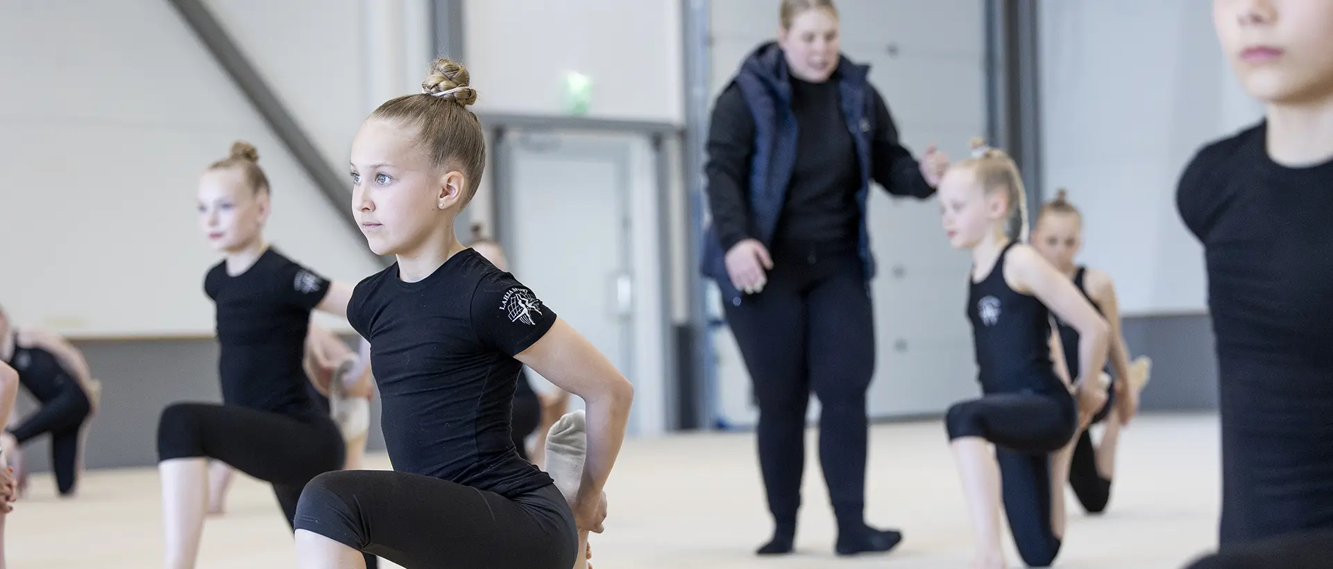 Voimistelijat venyttelevät treenisalissa. Valmentaja ohjeistaa urheilijoita taustalla.