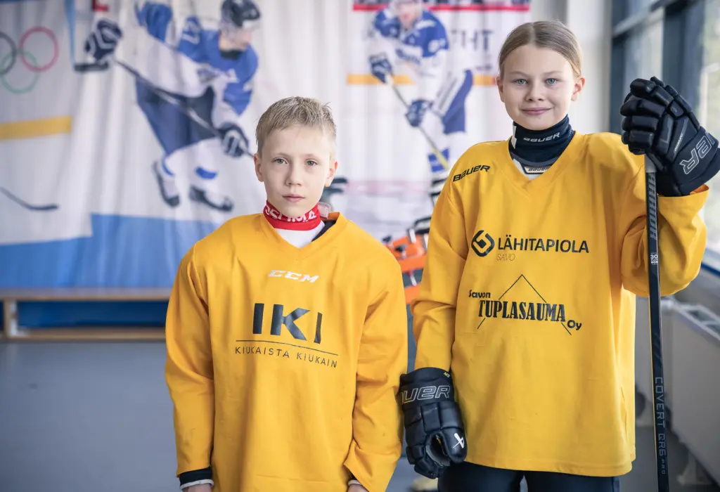 Kaksi nuorta jääkiekkoilijaa seisoo jäähallissa keltaisissa harjoituspaidoissa. Oikealla oleva pelaaja pitää jääkiekkomailaa kädessään.