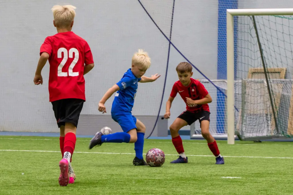Lapset pelaavat jalkapalloa sisähallissa. Siniseen peliasuun pukeutunut pelaaja kuljettaa palloa kahden punapukuisen pelaajan välissä.